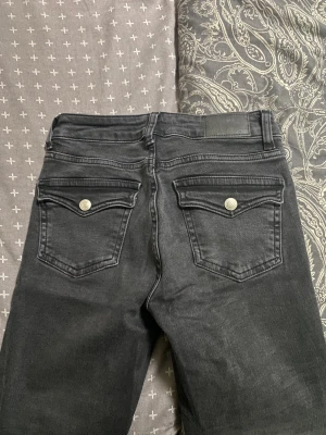 Svarta jeans från 157 med fickdetaljer - Säljer ett par svarta jeans från 157 med snygga ficklock och silverfärgade knappar bak. Jeansen har klassisk femficksdesign, raka ben och är tillverkade i mjukt denimtyg. Perfekta för dig som gillar en stilren look med lite edge.
