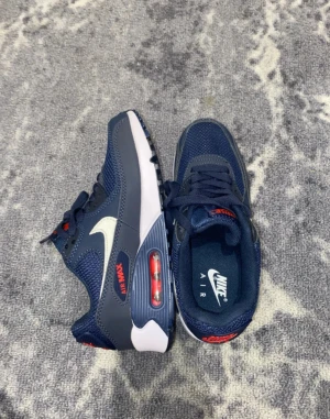 Nike Air Max 90 marinblå sneakers - Nike Air Max 90 sneakers i marinblå med vita och röda detaljer. Snygg mesh och syntet på ovandelen, klassisk Air Max-sula med synlig luftkudde och platt sula. Ikonisk Swoosh på sidan och Air Max-logga i rött. Perfekt för dig som gillar streetstyle.