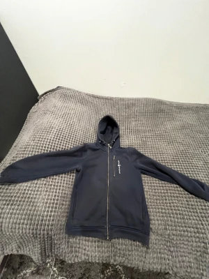 Mörkblå hoodie från Sail Racing - Snygg mörkblå hoodie från Sail Racing med dragkedja hela vägen och huva med snörning. Tröjan har en bröstficka med dragkedja och logga på vänster sida. Tillverkad i mjuk polyester, perfekt för chill eller när du vill vara bekväm. Lappaena inuti är borta för dom va lite jobbiga men annars är den i perfekt skick 