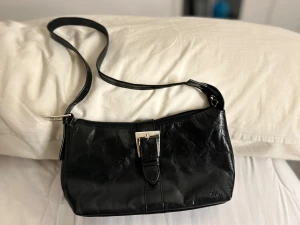 Svart axelväska från The Monte - Snygg svart axelväska i skinn från The Monte med justerbar rem och silvriga metalldetaljer. Väskan har ett spänne framtill, dragkedja upptill och flera fack inuti med beige foder. Perfekt storlek för det viktigaste och stilren look.