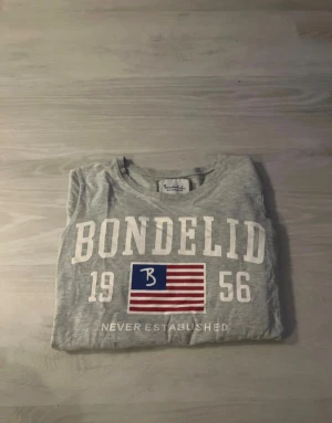 Grå tröja från Bondelid med tryck - Snygg grå långsarmad tröja från Bondelid i bomull med stort vitt tryck framtill, amerikansk flagga med blått och rött, samt texten 'Bondelid 1956'. Klassisk rund hals. Perfekt för dig som gillar stilrena och sportiga plagg.