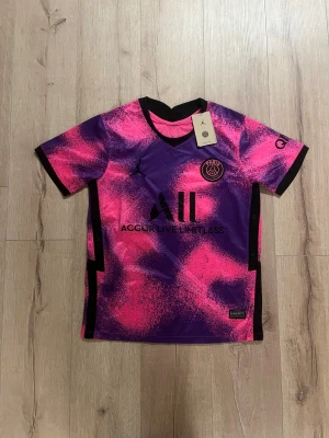 PSG Mbappé kortärmad fotbollströja S - Säljer en PSG fotbollströja med Mbappé #7 på ryggen, designad av Jordan. Tröjan är kortärmad och har ett unikt mönster i lila och rosa med svarta detaljer. Materialet är lätt och andas, perfekt för match eller träning. PSG-logga och sponsortryck på bröstet.