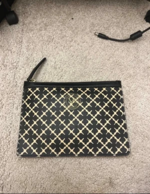  kuvertväska från Malene Birger - Kuvertväska från Malene Birger i svart och beige med grafiskt mönster och guldfärgad logga framtill. Väskan är i skinn och har dragkedja upptill. Perfekt storlek för det viktigaste och snygg att bära i handen.