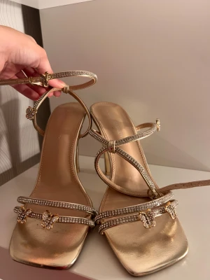 Guldiga sandaler med strass och fjärilar - Säljer ett par guldiga klackar från Kanada, köpta i butik för 1 år sen för ca 750 svenska kronor har bara använt 3-4 gånger. Skriv om ni vill ha specifika bilder eller om ni har frågor. Pris kan diskuteras!💕