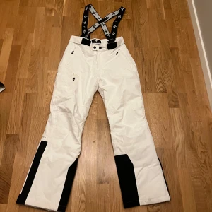 Vita täckbyxor Cross Sportswear S - Snygga vita täckbyxor från Cross Sportswear i storlek S med svarta detaljer på benen och justerbara hängslen med logga. Även avtagbara. Byxorna har dragkedjor vid fickorna och är perfekta för vinterns äventyr. Materialet är slitstarkt och vattenavvisande syntet.