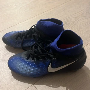 Nike Magista blå fotbollsskor - Nike Magista fotbollsskor i blått och svart med högt skaft och snörning. Skorna har en texturerad ovandel för bättre bollkontroll och en vit Nike-logga på sidan. Tillverkade i syntetmaterial med platt sula och rund tå, perfekta för dig som vill sticka ut på planen.