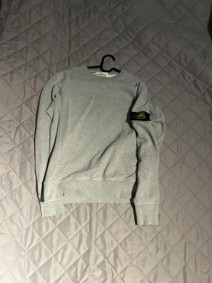 Grå sweatshirt från Stone Island - Säljer en stilren grå sweatshirt från Stone Island med klassisk rund hals och ribbade muddar. Tröjan har den ikoniska Stone Island-patchen på vänster ärm och är tillverkad i mjuk bomull. Perfekt för dig som gillar clean streetwear och vill ha något som funkar året runt.