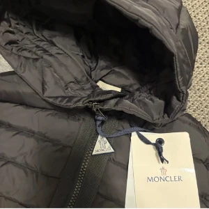 Svart hybridjacka från Moncler - Snygg svart hybridjacka från Moncler med quiltad pufferfront och huva i blankt material samt stickade ärmar. Jackan har dragkedja framtill, två fickor med tryckknappar och Moncler-logga på ärmen. Perfekt för höst  och vinter när du vill hålla stilen.