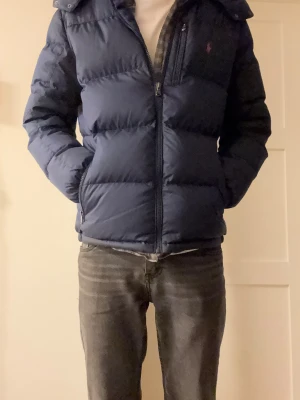 Mörkblå dunjacka från Polo Ralph Lauren - Snygg mörkblå dunjacka från Polo Ralph Lauren med huva och dragkedja. Jackan har klassisk pufferstil, tre fickor med dragkedja och ikonisk röd logga på bröstet. Perfekt för kalla dagar och riktigt skön att bära. Han på bilden är 180cm och väger 65kg. Finns inga deffekter på jackan. Den är i storlek 18-20 år XL, vilket motsvarar S.