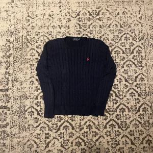 Mörkblå stickad tröja Polo Ralph Lauren - Kabelstickad mörkblå tröja från Polo Ralph Lauren i storlek M. Klassisk modell med rund halsringning och röd broderad logga på bröstet. Perfekt för dig som gillar tidlös och stilren design.