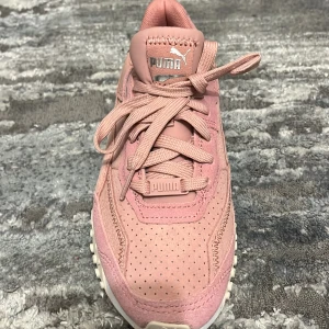 Rosa sneakers från Puma, strl 36 - Snygga rosa sneakers från Puma i storlek 36. Skorna har en perforerad ovandel i mocka och syntet, klassisk snörning och vit sula med mönstrat grepp. Ikonisk Puma-logga på både plös och häl. Perfekta för dig som gillar sportig och trendig stil. Dem är helt nya och oanvända. 