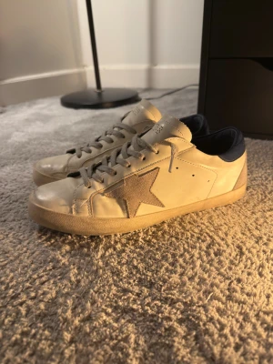 Vita Golden Goose sneakers - Snygga beige sneakers med grå snörning och en stor stjärna i mocka på sidan. Skorna har mörkblå detalj vid hälen och är tillverkade i skinn med inslag av mocka. Perfekta för en avslappnad och trendig look.