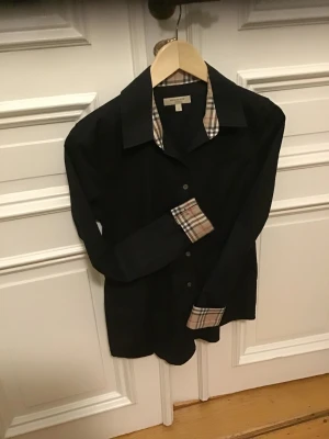 Svart Burberry skjorta med rutiga detaljer - Svart skjorta från Burberry med klassiska rutiga detaljer på krage och ärmslut. Skjortan har knappar framtill och lång ärm. Snygg och tidlös design som ger en clean look med en touch av Burberrys ikoniska mönster.