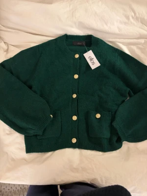 Grön kofta från Ellos med guldknappar - Mysig mörkgrön kofta från Ellos med rund hals och snygga guldknappar framtill. Koftan har två praktiska fickor och ribbade muddar vid ärmslut och nederkant. Perfekt för att addera lite färg till din garderob. 