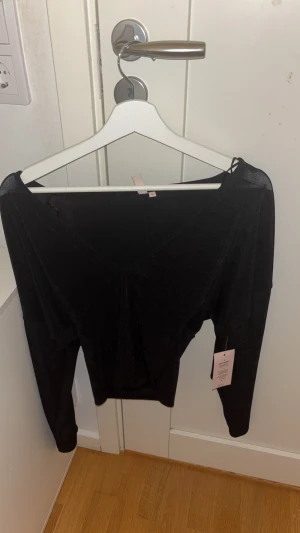 Svart  långärmad topp S - Svart  långärmad topp i storlek S med snygg v-ringning och lätt omlott-effekt framtill. Materialet har en skimrande finish som ger en cool look. Perfekt för dig som gillar att sticka ut med stilrena detaljer och glans.