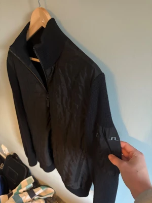 Svart J.Lindeberg Cardigan  - Snygg svart jacka från J.Lindeberg med quiltad front och stickade ärmar och rygg. Jackan har dragkedja framtill, hög krage och diskret JL-logga på ärmen. Perfekt för dig som vill ha en stilren och sportig look med både komfort och edge. I ett mycket fint skick, endast en liten skråma på JL märket men inget man tänker på annars. Storlek M👍 Bara att fråga om man undrar nåt.