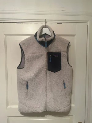 Vit fleeceväst från Patagonia - Nyskick, nypris: 1500kr. Mysig vit fleeceväst från Patagonia med blå detaljer och dragkedjor. Västen har en hög krage, tre fickor med dragkedja och en kontrasterande blå bröstficka. Perfekt för lager-på-lager och chill dagar.