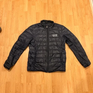 Svart pufferjacka från EA7 Armani - Snygg svart pufferjacka från EA7 Emporio Armani med quiltad design och diskret logga på bröstet. Jackan har dragkedja framtill och stårkrage, perfekt för kalla dagar. Materialet är lätt och syntetiskt, vilket gör den smidig att bära.