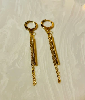 Guldiga örhängen med kedjor - Snygga guldiga örhängen med rund ring och hängande detaljer i form av tunna kedjor och spiralformade stavar. De har en modern och edgy look som verkligen sticker ut. Perfekta för dig som gillar statement-smycken och vill addera lite extra till din stil.