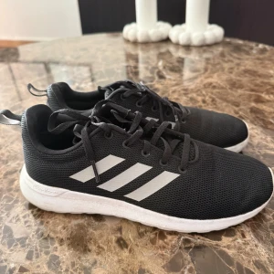 Svarta Adidas sneakers med vit sula - Snygga svarta Adidas sneakers med klassiska vita ränder på sidan och vit sula. Skorna har snörning och är tillverkade i meshmaterial för bra andningsförmåga. Perfekta för en sportig och avslappnad stil.
