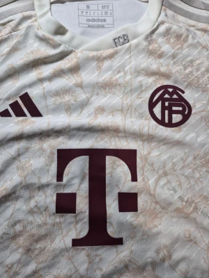 FC Bayern München blommig fotbollströja S - Säljer en FC Bayern München fotbollströja från Adidas i storlek S. Tröjan är vit med ljusbeige blommigt mönster, har korta ärmar och rund hals. Märken från Adidas och FCB syns framtill, samt ett stort T-tryck. Materialet är lätt och andas, perfekt för match eller träning.