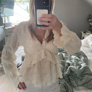 Krämvit blus med volang - Säljer en krämvit blus från Gina Tricot med v-ringning och långa ärmar. Blusen har ett strukturerat tyg och volangdetaljer framtill och baktill som ger en romantisk känsla. Perfekt att styla med jeans eller kjol.