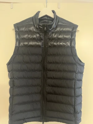 Svart dunväst från J.Lindeberg - Snygg svart dunväst från J.Lindeberg med quiltad design och hög krage. Västen har en stilren look med diskret logga på bröstet och dragkedja framtill. Perfekt att slänga över en hoodie eller tröja för extra värme. Fickor med dragkedja på sidorna. Använd fåtal gånger! Som ny