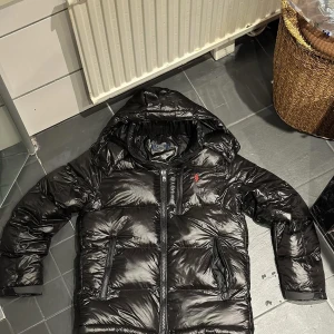 Svart pufferjacka från Polo Ralph Lauren - Svart pufferjacka från Polo Ralph Lauren med glansig finish, huva och dragkedja framtill. Jackan har två stora fickor med dragkedja och en röd Polo-logga på bröstet. Perfekt för kalla dagar med sin tjocka vaddering och stilrena look. Pris kan diskuteras vid snabb köp