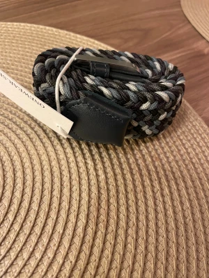 Flätat bälte från ONEWear Sweden - Snyggt flätat bälte från ONEWear Sweden i svart, grått och vitt. Bältet har detaljer i svart skinn och är gjort i ett elastiskt, vävt material som ger en cool och modern look. Perfekt accessoar för att lyfta din outfit. Helt ny aldrig använd.
