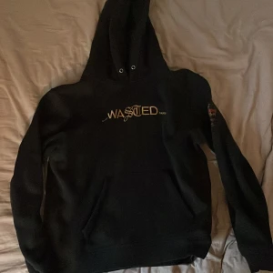 Svart hoodie från Wasted Paris - Svart hoodie från Wasted Paris med broderad logga i guld och vitt på bröstet. På ena ärmen finns en patch med röd och gul grafik och texten 'Under Influence SS21'. Tröjan har huva med snörning och en stor magficka framtill. Perfekt för dig som gillar streetwear. Oversized. Hör av er vid frågor