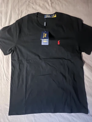 Svart t-shirt från Polo Ralph Lauren - Svart t-shirt från Polo Ralph Lauren med klassisk rund hals och korta ärmar. På bröstet finns den ikoniska röda Polo-loggan broderad. T-shirten är gjord i mjuk bomull och har en clean, stilren look som funkar till allt.