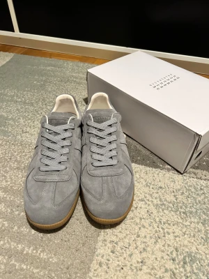 Maison Margiela Gats - Maison Margiela sneakers i grå mocka med klassisk rund tå och platt gummisula i beige. Skorna har snörning och vit insida, samt diskret logga på tungan och undersidan. Snygg minimalistisk design som passar till streetwear och casual outfits.