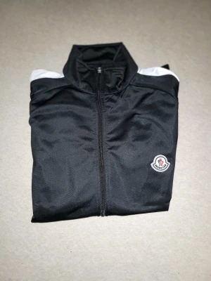 Svart Moncler vindjacka med vita detaljer - Svart vindjacka från Moncler med vita ränder längs axlarna och ärmarna. Jackan har dragkedja framtill, och Moncler-logga på bröstet. Tillverkad i ett slätt syntetmaterial.