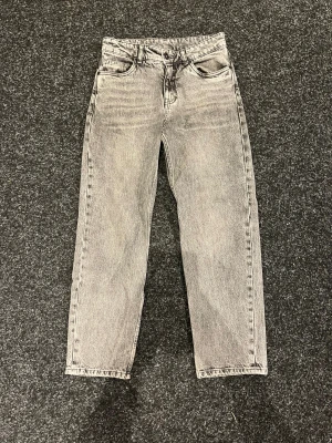 Grå jeans från Kappahl, storlek 152 - Säljer ett par grå jeans från Kappahl i storlek 152. De har en snygg tvättad look, klassisk femficksdesign och raka ben. Jeansen är tillverkade i jeansmaterial och har knapp och dragkedja framtill. Perfekt för dig som gillar en chill och stilren vibe.