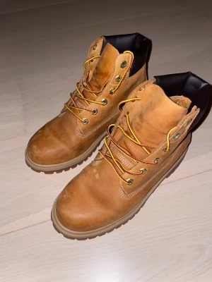 Timberland boots i mocka, brun - Klassiska Timberland boots i ljusbrun mocka med robust gummisula och gul-röda snören. Skorna har rund tå, kraftig sula och vadderad svart kant upptill för extra komfort. Perfekta för höst och vinter, ikonisk streetstil.