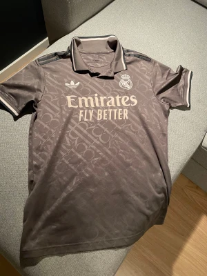 Real Madrid x Adidas fotbollströja M - Snygg mörkgrå Real Madrid fotbollströja från Adidas med diskret mönster, klassisk krage och korta ärmar. Tröjan har broderat klubbmärke, Adidas-logga och Emirates Fly Better-tryck på bröstet. Tillverkad i lätt och ventilerande polyester.
