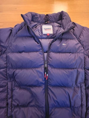 Mörkblå pufferjacka Tommy Jeans - Snygg mörkblå pufferjacka från Tommy Jeans med klassisk logga på bröstet och ärmen. Jackan har dragkedja med röda och vita detaljer, hög krage och två sidofickor. Perfekt för kalla dagar. Knappt några spår på användning. Orginalpris 2400kr.