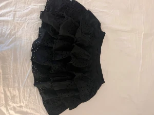 Svart volangkjol med broderade hål - Supersöt svart kortkjol med flera lager volanger och broderade hål för en extra cool look. Kjolen har resår i midjan och är perfekt för dig som gillar en lekfull och trendig stil. Materialet känns lätt och luftig.