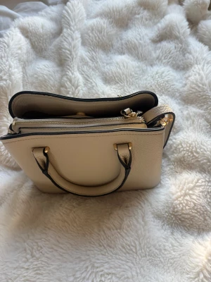 Beige handväska med gulddetaljer - Snygg beige handväska med guldfärgade metalldetaljer. Väskan har både handtag och justerbar axelrem, samt flera fack och dragkedja upptill. Perfekt storlek för att få plats med allt möjligt. Köpt utomlands förra sommaren, men knappt aldrig använt. 