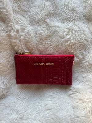 Röd plånbok Michael Kors med reptilmönster - Snygg röd plånbok från Michael Kors med reptilmönster och guldfärgade dragkedjor. Plånboken har flera fack och är perfekt för kort och kontanter. Tidigare köpt second hand. 