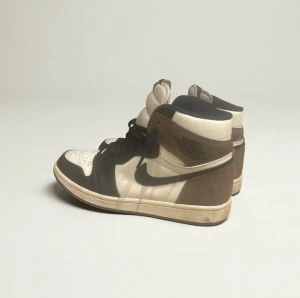 Jordan 1 High Mocha - Nike Air Jordan 1 High Mocha med svart swoosh, bruna och vita paneler samt svarta snören. Skorna har en klassisk high-top siluett, rund tå och platt sula. Materialet är en mix av skinn och mocka, vilket ger en lyxig känsla. Perfekt för dig som gillar streetwear och sneakers med edge.