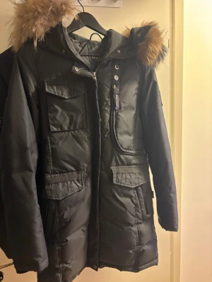 Svart parkasjacka med päls från Hollies - Svart parkasjacka från Hollies med stor huva och fluffig päls i bruna toner. Jackan har flera fickor med lock, dragkedja och knappar framtill. Långärmad och quiltad design som håller dig varm under kalla dagar. Perfekt för dig som gillar stilren och funktionell vinterjacka.