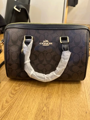 Brun handväska  - Brun väska med klassiskt mönster och guldiga detaljer. Helt ny och oanvänd. Storlek på väskan 27x14x19 cm. Inte äkta! Pris kan diskuteras. 