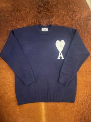 Marinblå stickad tröja från Ami - Snygg marinblå stickad tröja från Ami med vitt hjärta och bokstaven A broderat på bröstet. Tröjan har rund hals, ribbade muddar och långärmad passform. Perfekt för dig som gillar stilrena detaljer och vill ha något som sticker ut lite extra.