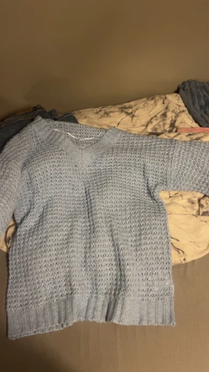 Ljusblå stickad v-ringad tröja - Säljer en ljusblå stickad tröja med v-ringad hals och långärmad design. Tröjan har ett snyggt hålmönster och breda ribbade muddar vid ärmslut och nederkant. Perfekt för dig som gillar mysiga och stilrena plagg.