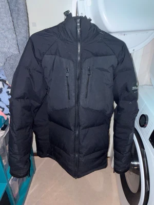 Svart Sail Racing dunjacka unisex - Svart dunjacka från Sail Racing, modell Antarctica Expedition. Jackan har hög krage, två bröstfickor med dragkedja och broderad logga på bröstet. Materialet är slitstarkt och jackan är vadderad för extra värme. Perfekt för kalla vinterdagar.