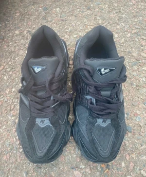 New Balance 9060 svarta sneakers - Säljer ett par New Balance 9060 sneakers i helsvart färg med chunky sula och coola detaljer. Skorna har mesh och mocka på ovandelen, snörning och en blå detalj på hälen. Perfekta för dig som gillar streetwear och vill sticka ut.