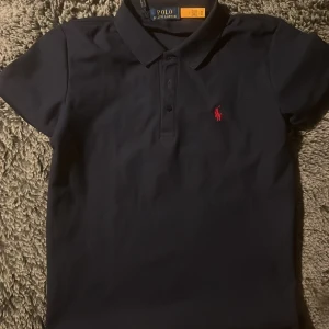 Mörkblå pikétröja från Polo Ralph Lauren - Klassisk mörkblå pikétröja från Polo Ralph Lauren med röd broderad logga på bröstet. Tröjan har korta ärmar, krage och knappar framtill. Perfekt för en stilren och sportig look.
