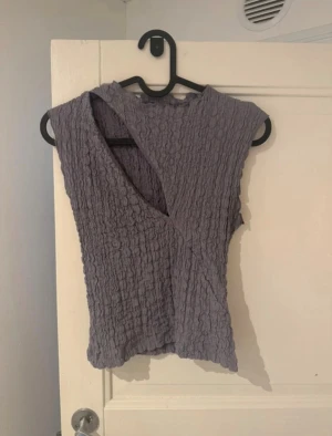 Lila omlott topp med struktur - Säljer en lila omlott topp med coolt strukturerat tyg och v-ringad hals. Toppen är ärmlös och har en unik omlott-design framtill som ger en snygg och modern look. Perfekt att styla med jeans eller kjol för en trendig vibe.