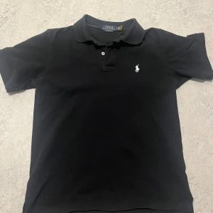 Svart pikétröja från Ralph Lauren - Svart pikétröja från Polo Ralph Lauren med klassisk krage och två knappar framtill. Tröjan har det ikoniska broderade logot i vitt på bröstet. Materialet är mjuk bomull och modellen är slim fit för en snygg siluett. Byten funkar.